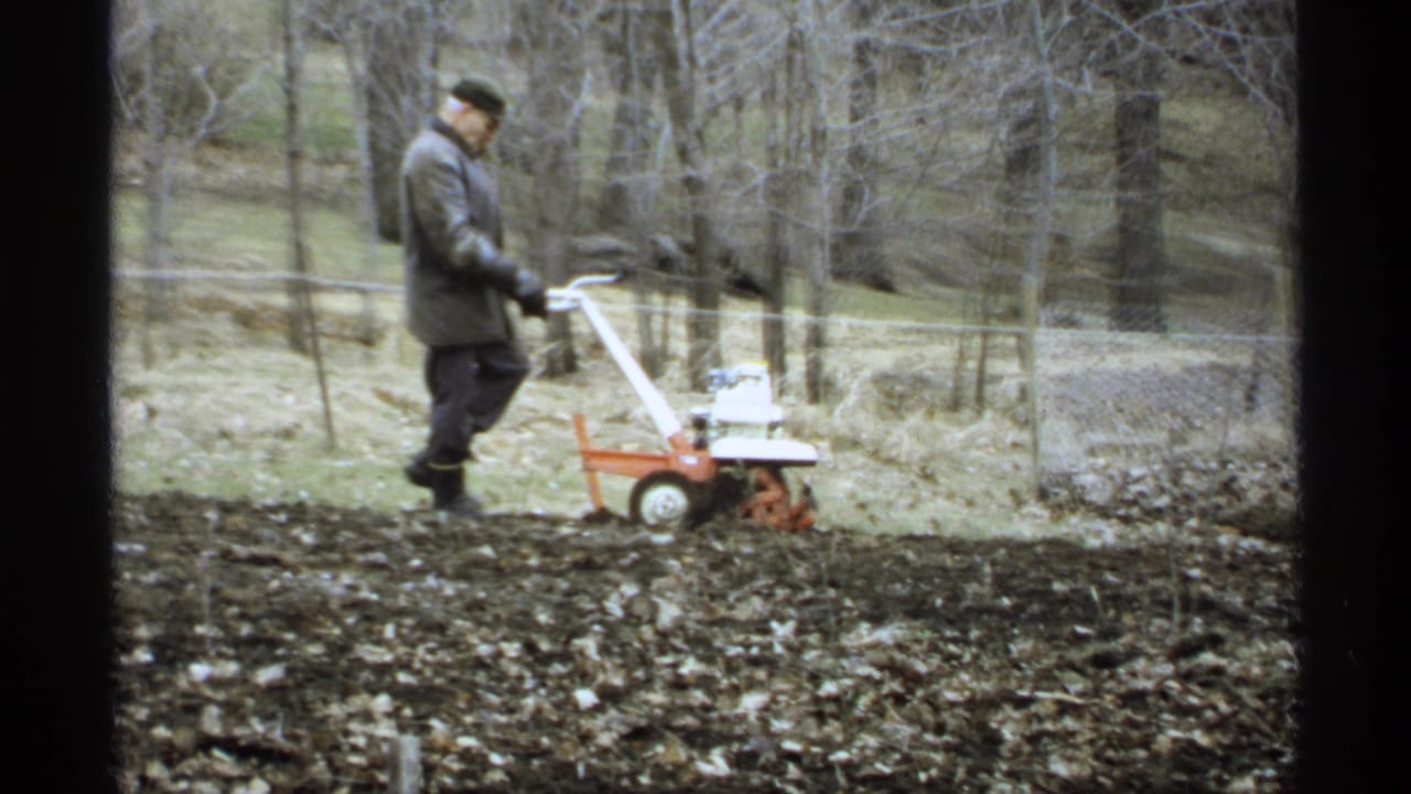 Man in a grey coat pushes a small tiller across dark garden soil near a wire fence.