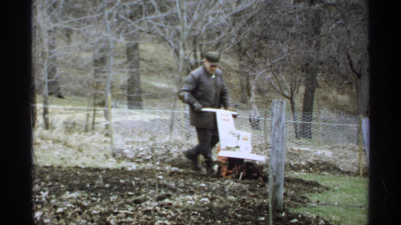 Man guides an orange and white tiller beside a wire fence and garden post.