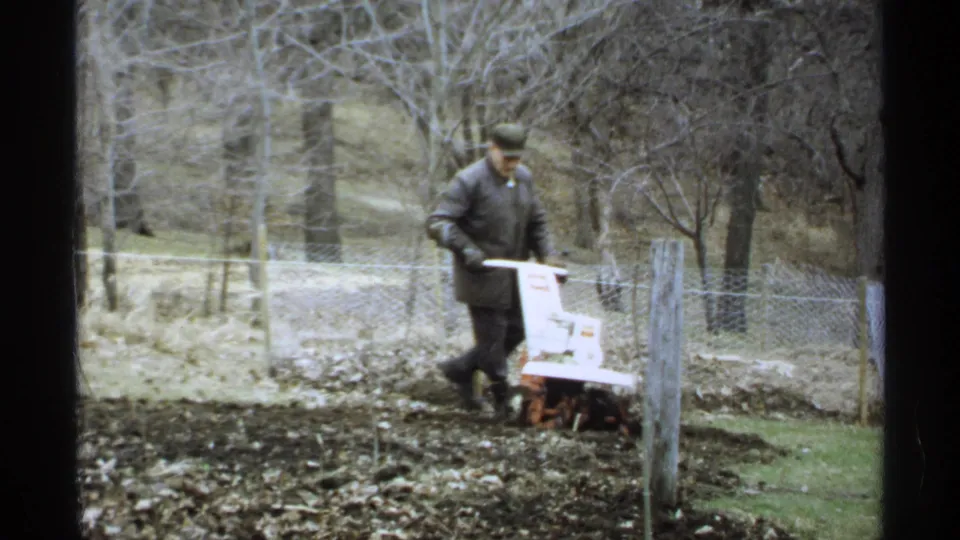 The man continues tilling a backyard garden in La Crosse, with wire fencing, bare trees, and muted 8mm color visible.