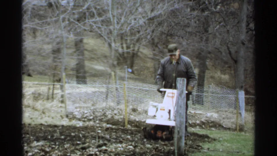 The man works the small tiller near a fence post, with bare trees and cloudy La Crosse yard scenery behind him.