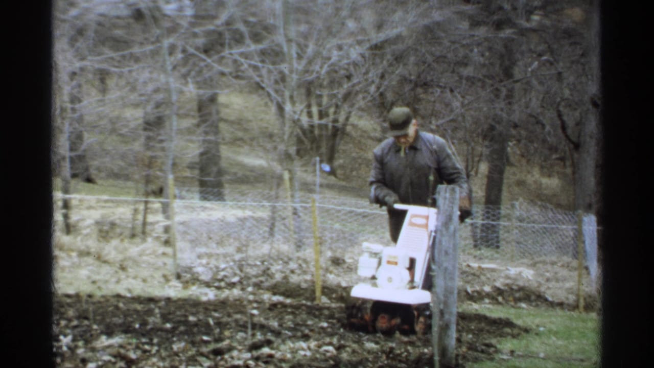 In 1971 La Crosse, Wisconsin home-movie footage, a man guides a white tiller through dark garden soil near bare trees and a fence.