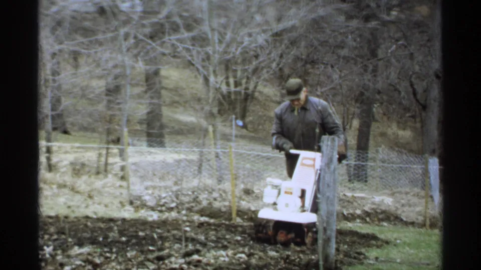 In 1971 La Crosse, Wisconsin home-movie footage, a man guides a white tiller through dark garden soil near bare trees and a fence.