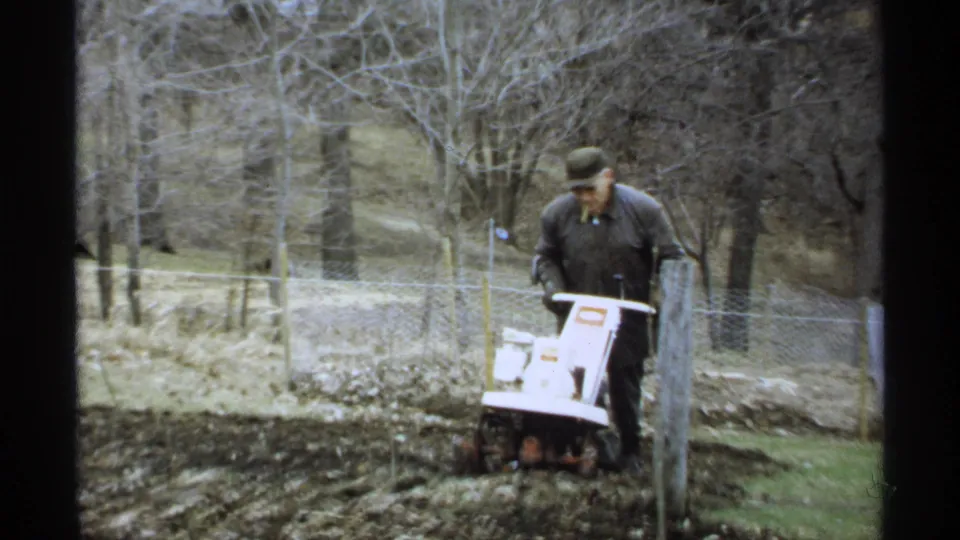 The man continues tilling a garden plot in a wooded La Crosse yard, with wire fencing and bare trees visible behind him.