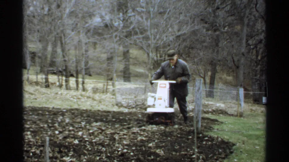 The 8mm frame shows the man steering the tiller across the garden bed, framed by trees, grass, and a wire fence.
