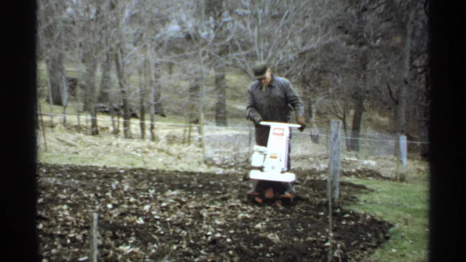 At the end of the clip, the man faces forward while guiding the white tiller through a garden plot in the wooded Wisconsin yard.