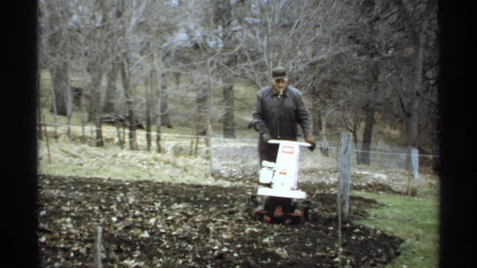 In La Crosse, Wisconsin, in 1971, a man guides a white motorized tiller through dark soil near bare trees and a wire fence.