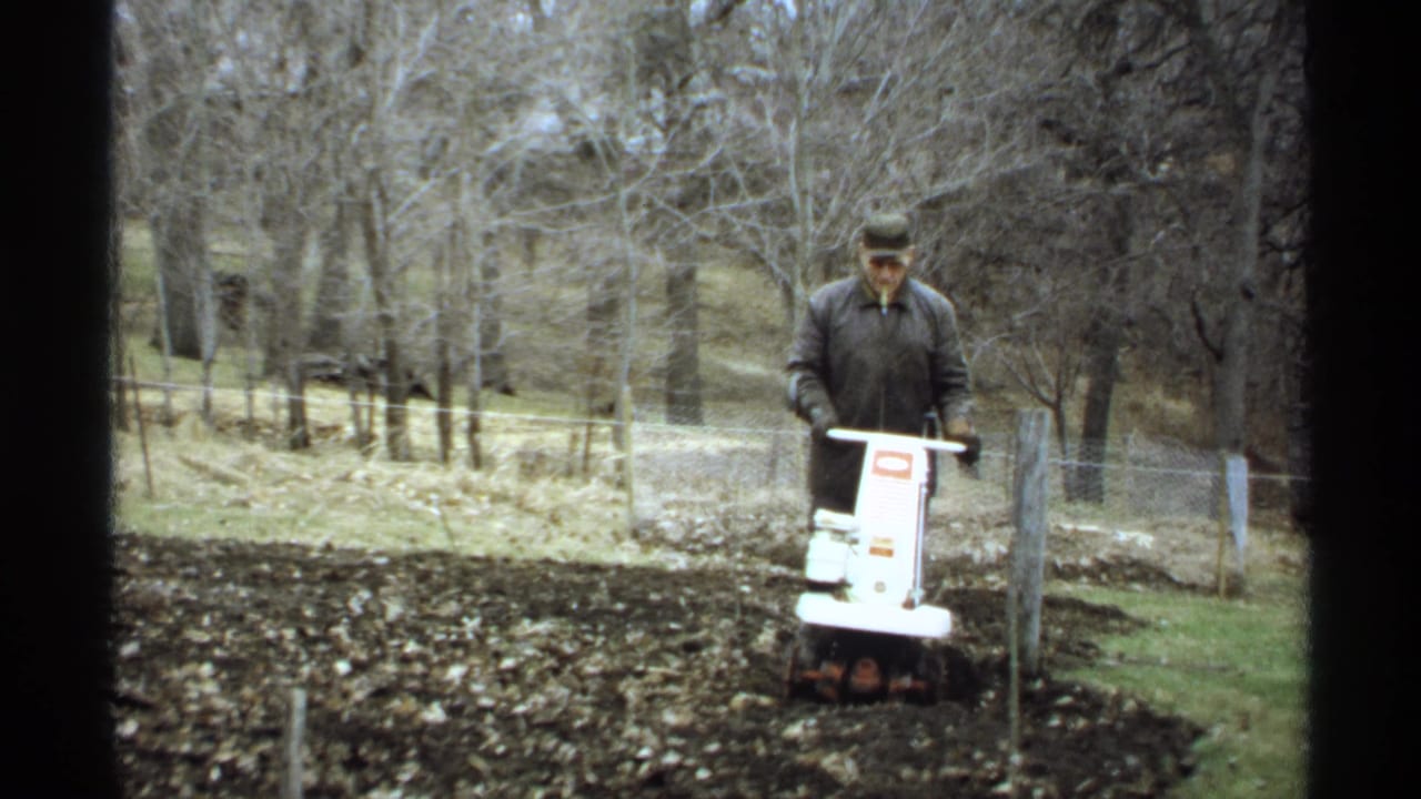 The 1971 La Crosse home-movie frame shows a man working soil with a white tiller in a quiet outdoor yard setting.