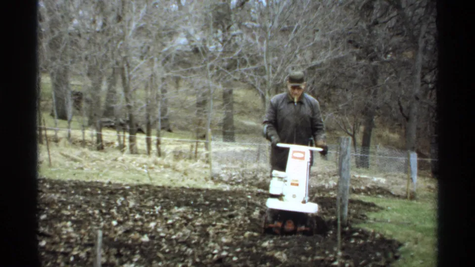 The 1971 La Crosse home-movie frame shows a man working soil with a white tiller in a quiet outdoor yard setting.