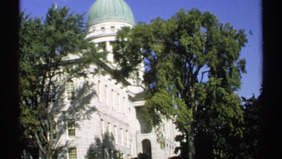 Low-angle 1971 home-movie view of the Maine State Capitol facade and green dome through leafy trees.