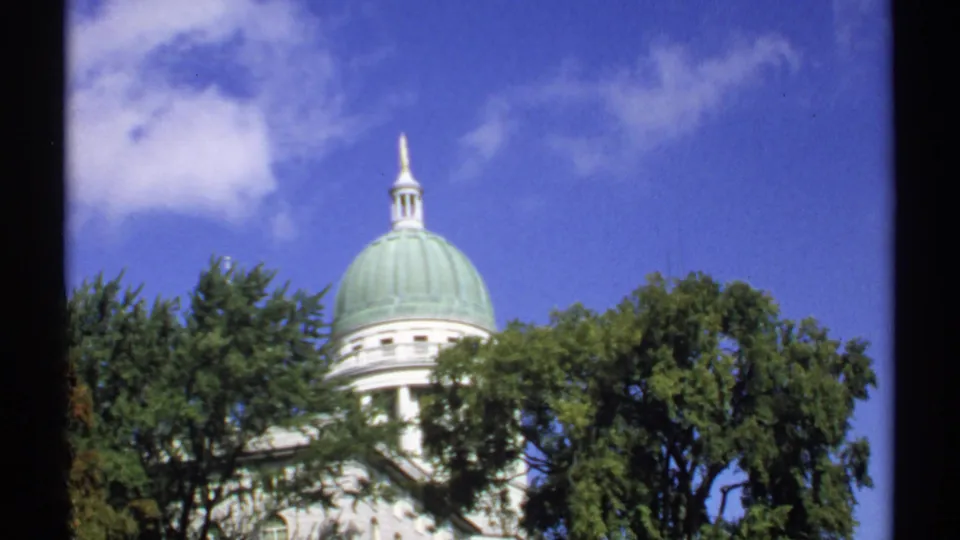 1971 Augusta view of the Maine State Capitol dome and spire rising above trees against a bright sky.