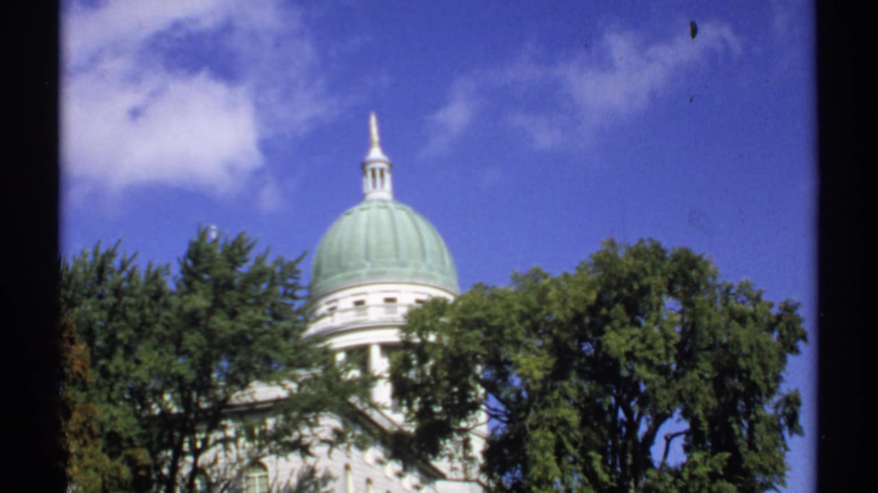 1971 Maine 8mm frame of a white government building's green dome rising above trees beneath a bright blue sky.