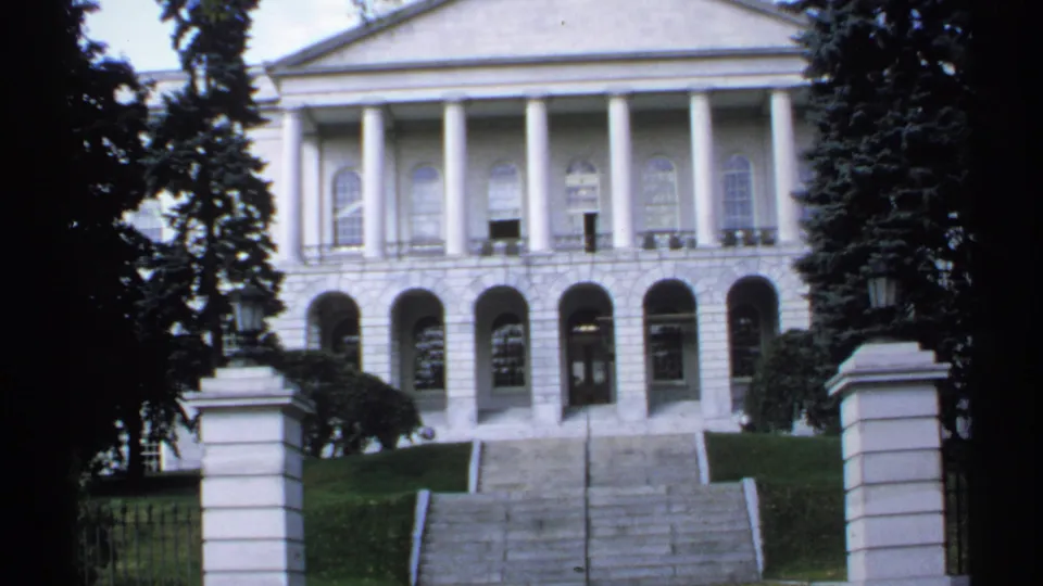 1971 Maine home-movie frame of a white classical government building facade, broad steps, lawn, and trees.