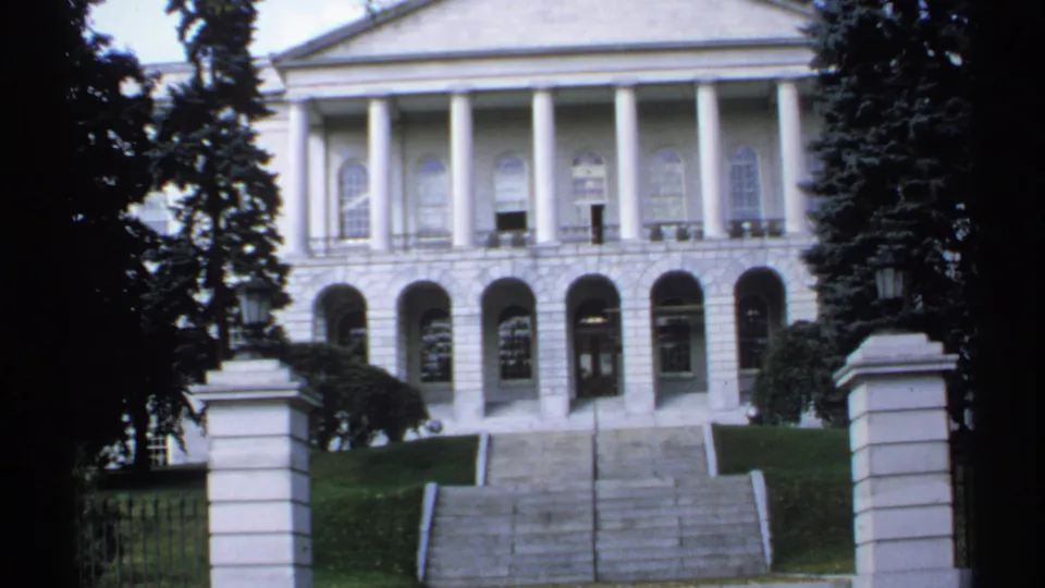 1971 Maine home-movie frame of a white classical civic building seen past gate posts, with columns, arched entries, steps, lawn, and evergreens.
