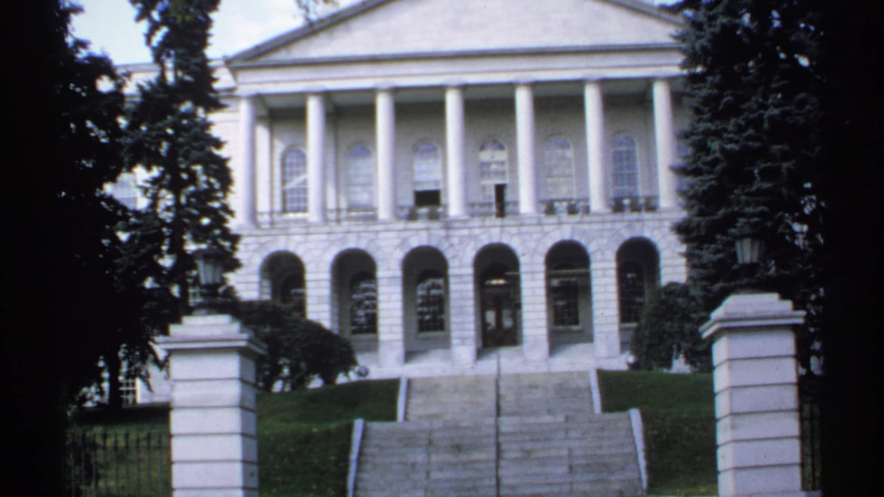 White columned building with arched lower level, steps, gate posts, lawn, and trees.