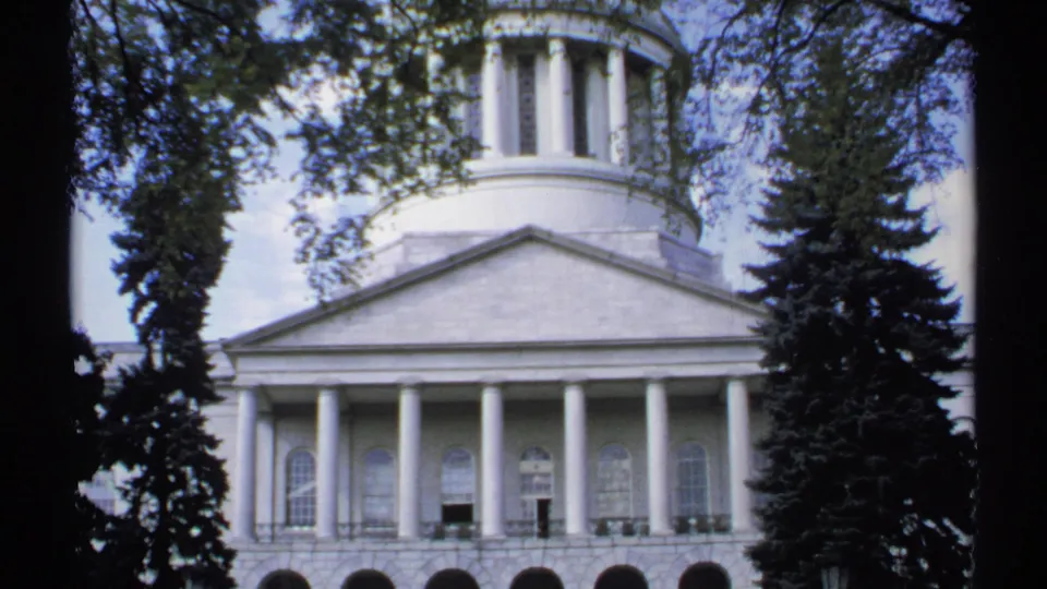1971 Maine frame showing the upper dome, triangular pediment, tall columns, arched windows, and shaded trees around a white civic building.
