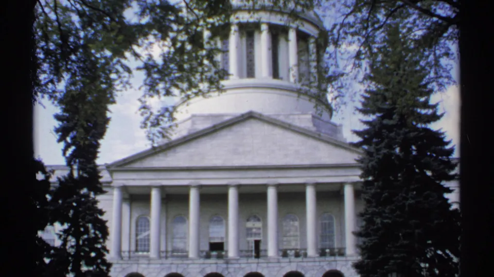 1971 Maine home-movie frame of a white domed civic structure, with columns and arched windows partly framed by dark trees.