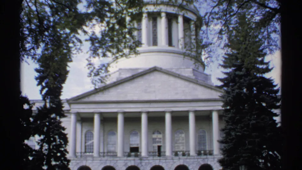 1971 Maine 8mm frame showing a white classical public building with columns and cupola framed by dark tree trunks.