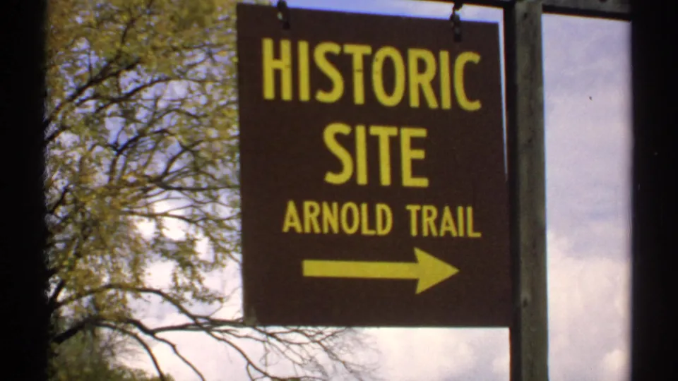 1971 Maine 8mm frame of the Arnold Trail historic-site sign, showing bold yellow lettering, arrow, signpost, leafy branches, and cloudy sky.