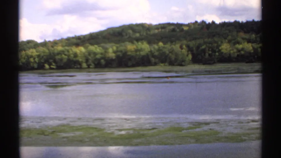 1971 Maine 8mm landscape view of a calm lake with green shoreline vegetation, forested hills, and a partly cloudy sky.
