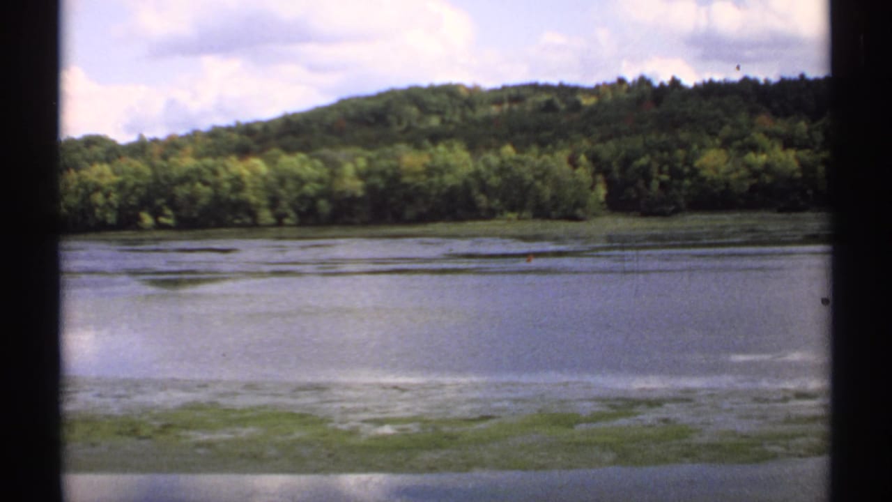 1971 Maine 8mm frame showing a calm lake, low green shoreline growth, wooded hill, and partly cloudy sky.