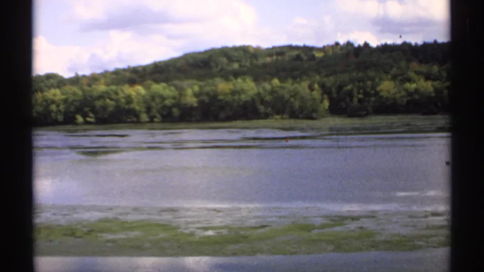 1971 Maine 8mm frame showing a calm lake, low green shoreline growth, wooded hill, and partly cloudy sky.