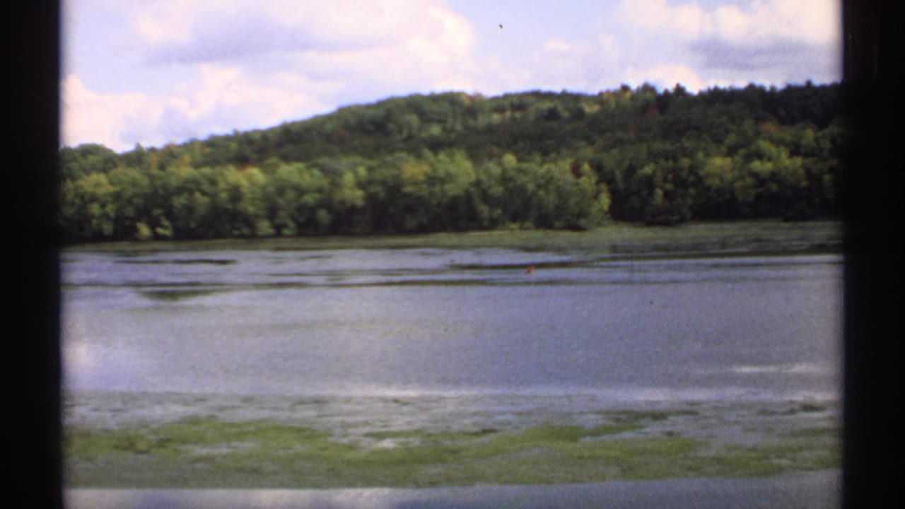 1971 Maine home-movie frame with still water, a forested ridge, cloudy sky, and visible 8mm frame borders.