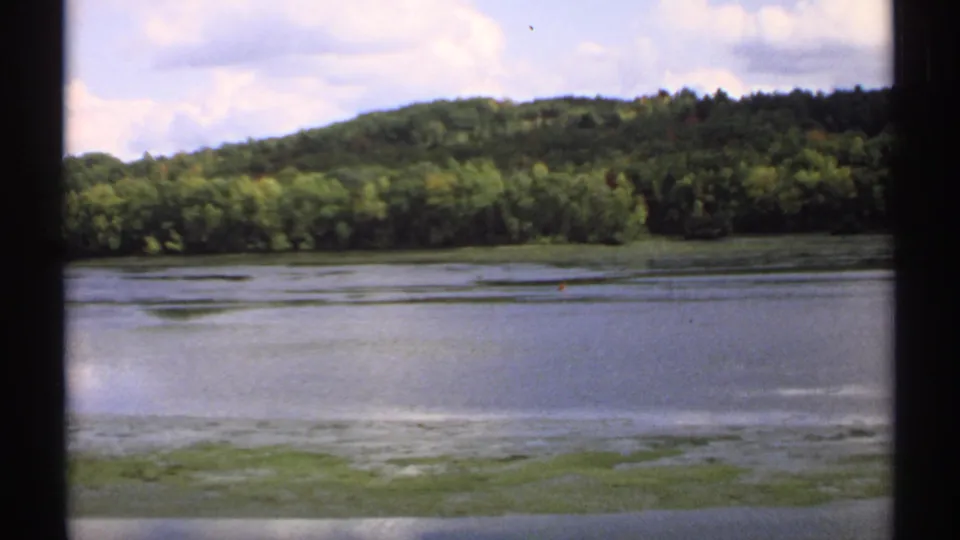 1971 Maine home-movie frame with still water, a forested ridge, cloudy sky, and visible 8mm frame borders.