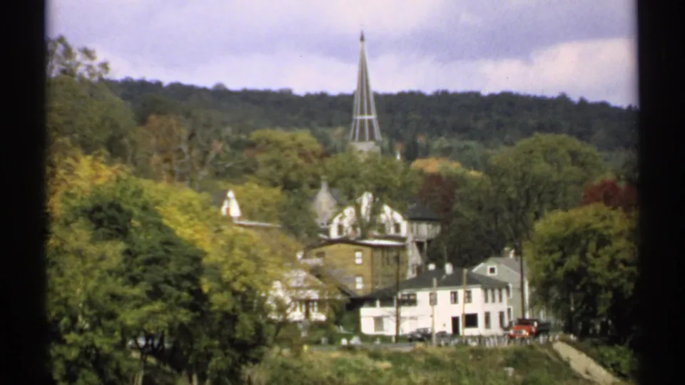 1971 Maine 8mm view of a small town set in trees, with clapboard houses, a tall church steeple, and wooded hills beyond.
