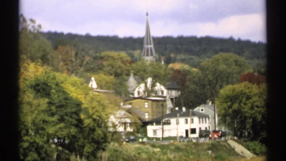 1971 Maine home-movie frame showing a tree-lined town, white houses, a pointed church steeple, and wooded hillside in the background.