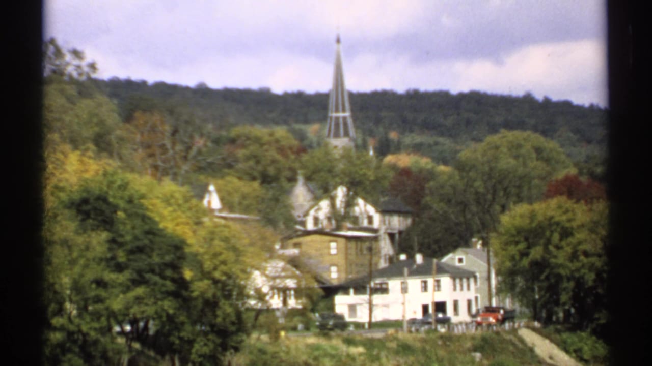 Similar hillside town view with steeple, houses, trees, and a road below.