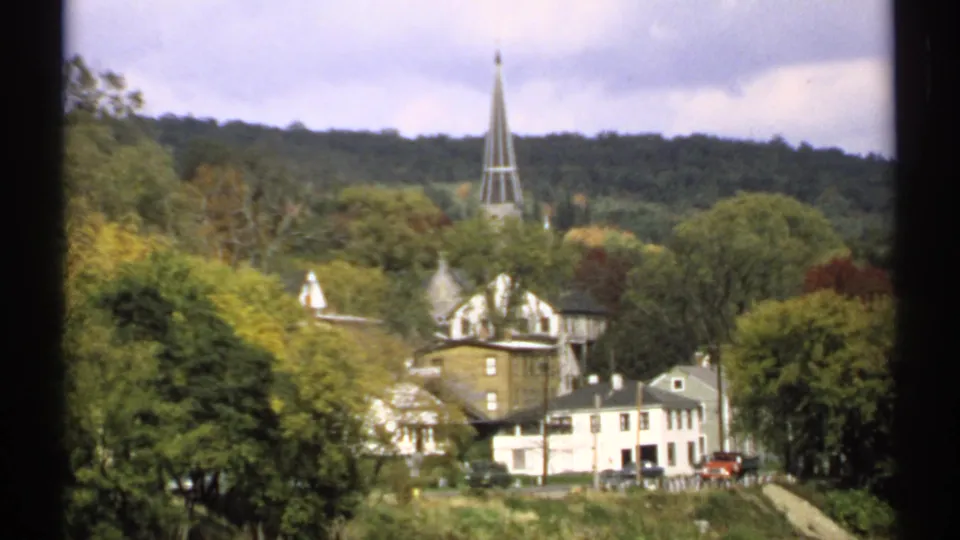 Small Maine townscape in 1971 with a tall steeple, white houses, trees, and a few parked cars along the road.