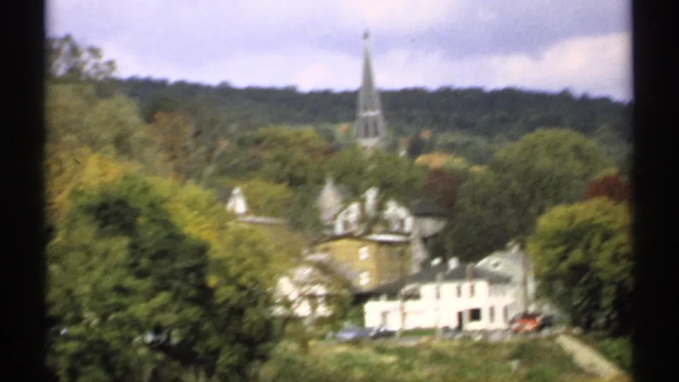 Motion-softened 1971 Maine town view from a moving vehicle, with steeple, hillside trees, houses, and roadside cars.