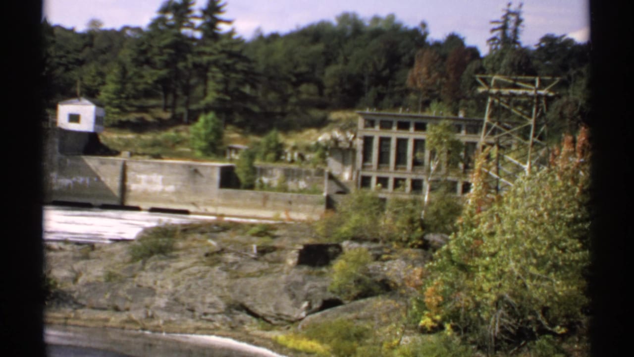 1971 Maine 8mm frame showing a concrete dam, small white utility building, transmission tower, trees, and rocky riverbank.
