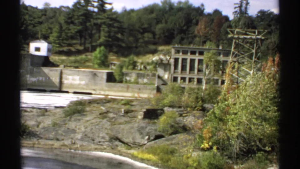 1971 Maine 8mm frame with a wide visitor view of the dam, white utility building, transmission structure, wooded hillside, and rocky foreground.