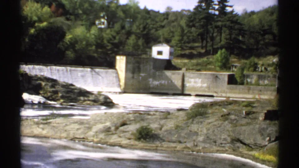 1971 Maine 8mm frame showing spillway water, concrete dam walls, a small white building, rocky riverbed, and surrounding trees.