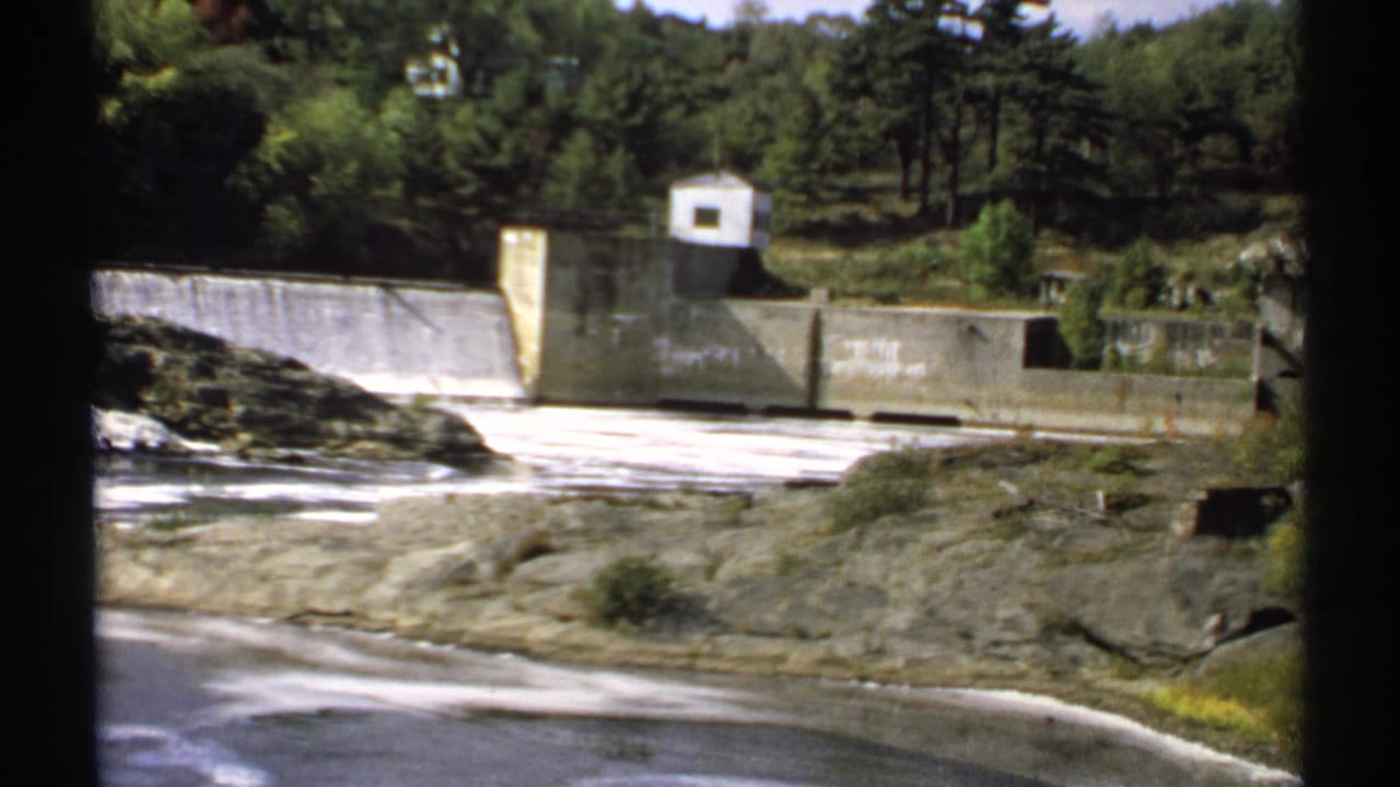 1971 Maine 8mm frame showing a concrete dam, spillway flow, rocky shoreline, trees, and a small white control building.