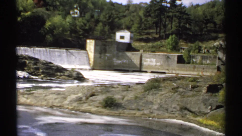 1971 Maine 8mm frame showing a concrete dam, spillway flow, rocky shoreline, trees, and a small white control building.