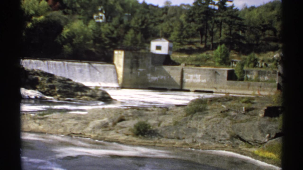 1971 Maine home-movie frame of a dam and reservoir edge, with white water, rock ledges, and dense trees behind the structure.