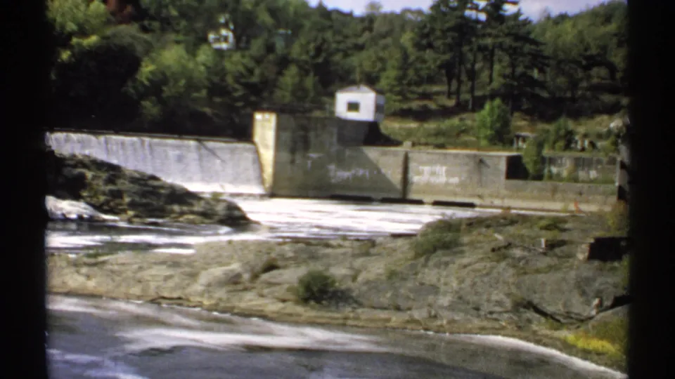 1971 Maine home-movie frame of a dam and reservoir edge, with white water, rock ledges, and dense trees behind the structure.