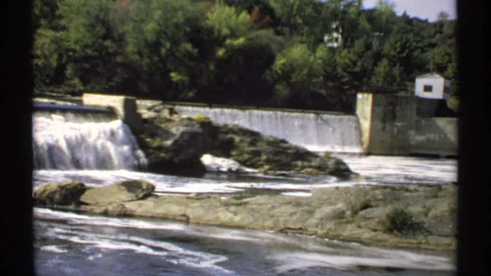 1971 Maine 8mm view of spillway water dropping beside rock ledges, with a concrete wall, small building, and trees in the background.