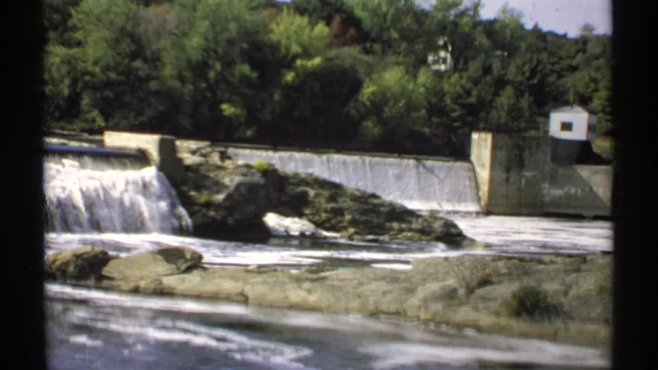 1971 Maine frame showing foamy water below a concrete spillway, rocky foreground, wooded hillside, and a small white control structure.