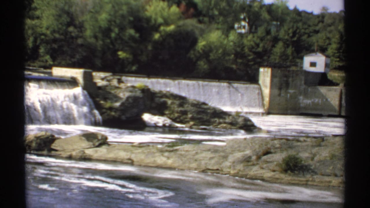 Concrete dam and low waterfall beside rocky riverbanks and trees.