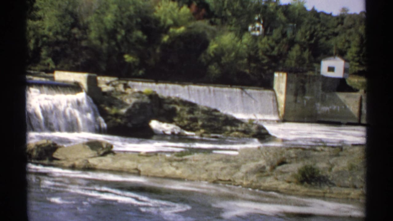 1971 Maine home-movie frame of water flowing over a concrete dam, with river rocks, green trees, and a small white structure at right.