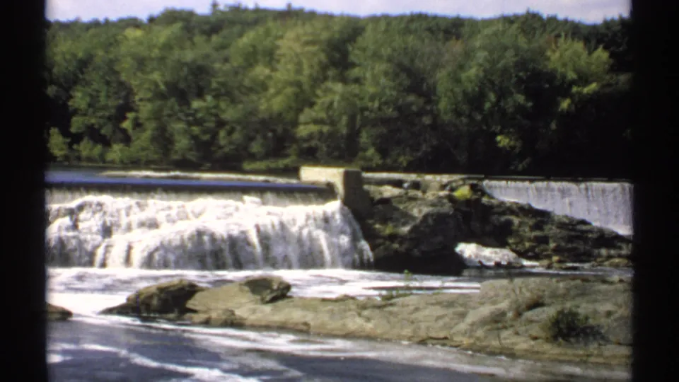 1971 Maine 8mm view of a low waterfall and dam across a rocky river, framed by dense green trees in the background.