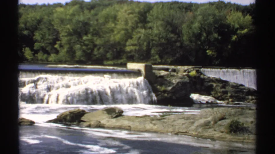 1971 Maine home-movie frame showing cascading dam water, exposed river rocks, foamy flow, and a wooded tree line beyond.