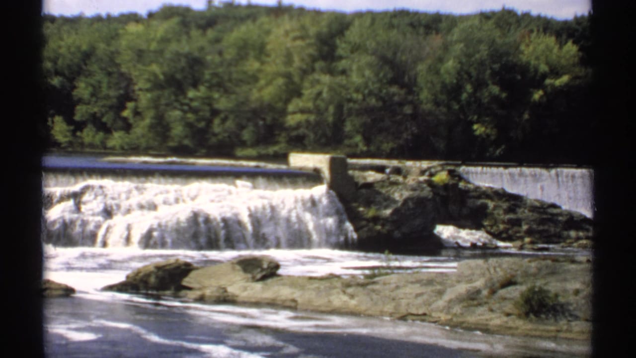 Wide river cascading over a low dam with rocks and trees beyond.