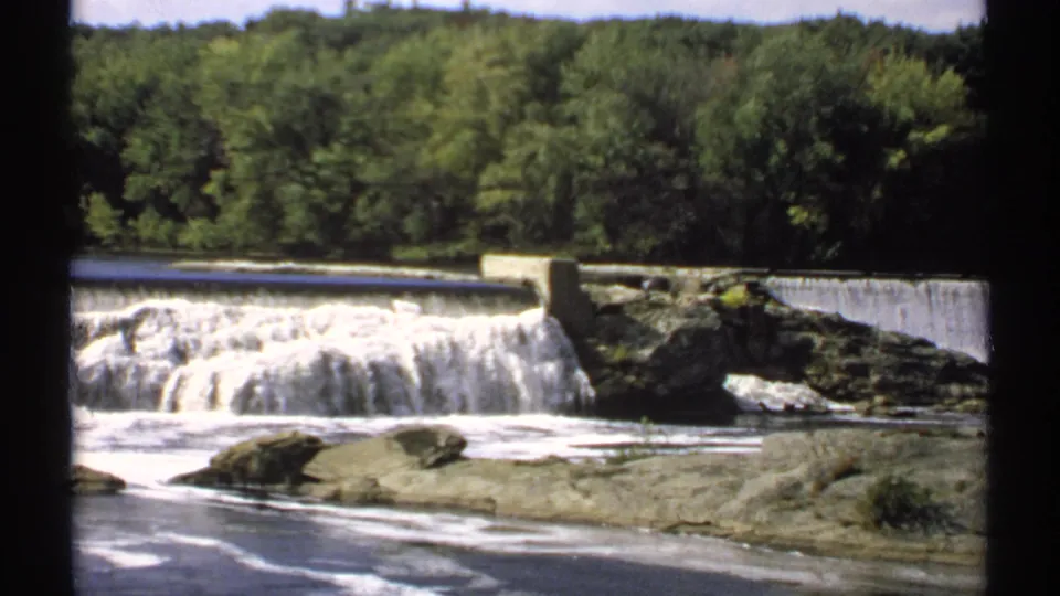 1971 Maine 8mm frame showing a wide river spilling over a low dam, with rocky foreground water and dense green trees behind.