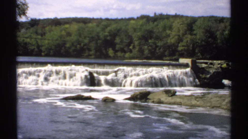 1971 Maine 8mm frame of white water pouring over a low dam, with rocks in the river and green forest across the bank.