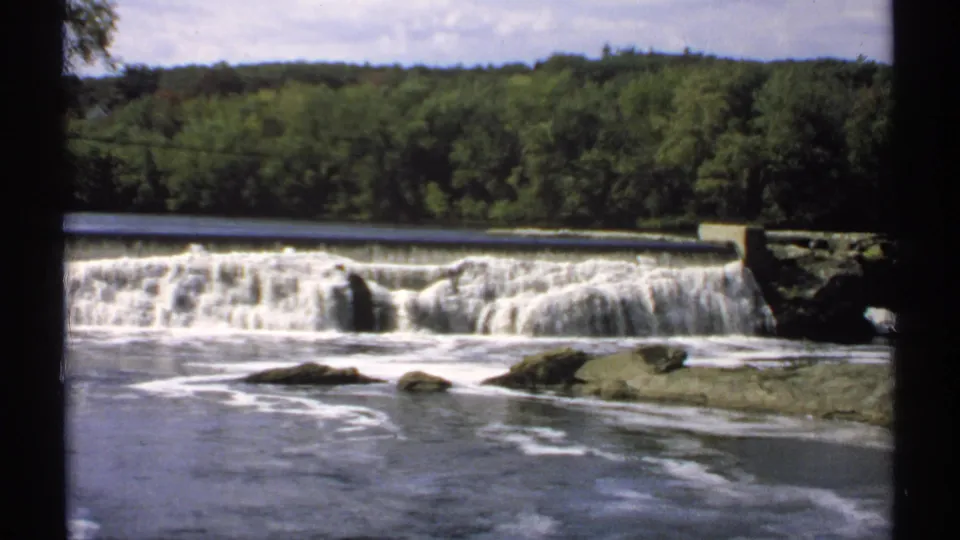 1971 Maine 8mm frame showing a wooded riverbank above a low dam, with foamy water moving around rocks in front.
