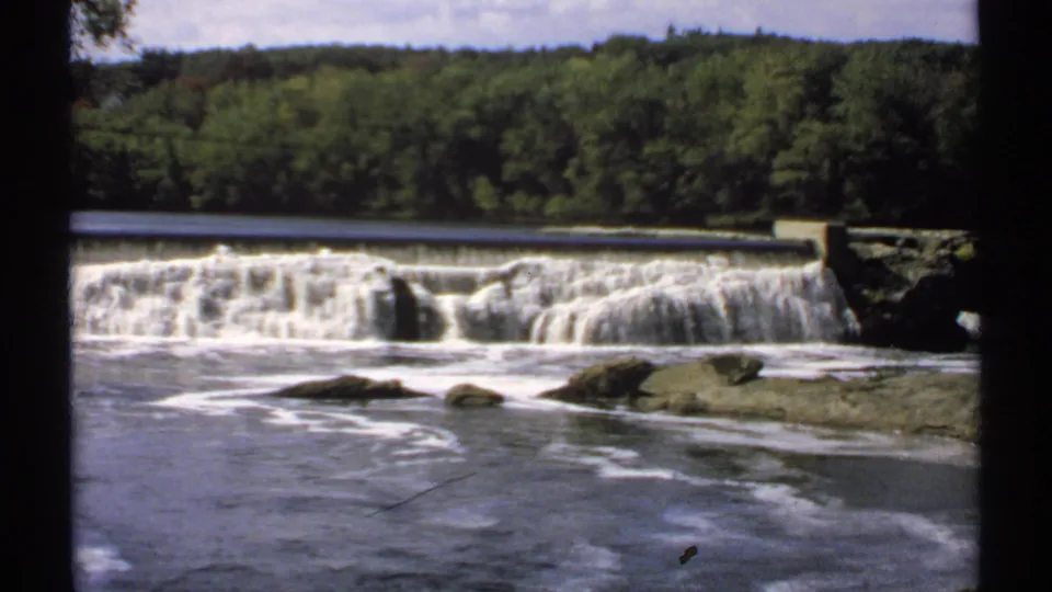 1971 Maine 8mm frame of a broad low dam behind river foam and exposed rocks, framed by dense green trees and film edges.